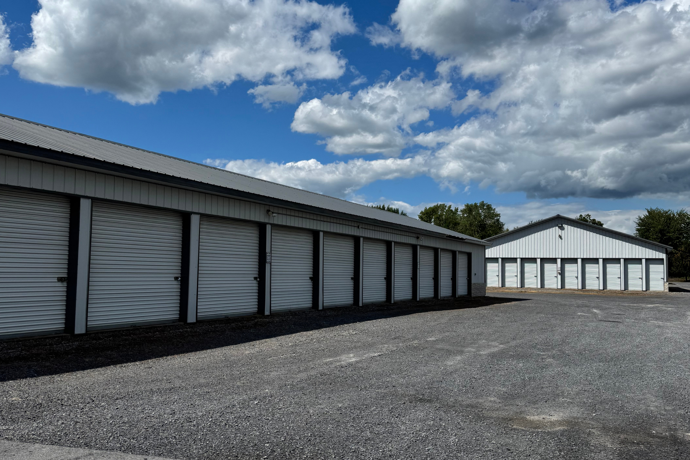 Close-up view of a self-storage building lined with white roll-up doors, with additional storage buildings visible in the background on a sunny day.